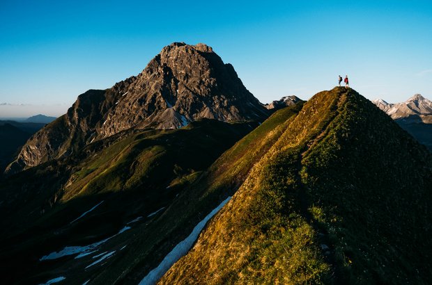 Lech Gipfel Zwei Personen gehen einen schmalen Pfad entlang und haben ein schönes Bergpanorama um sich.